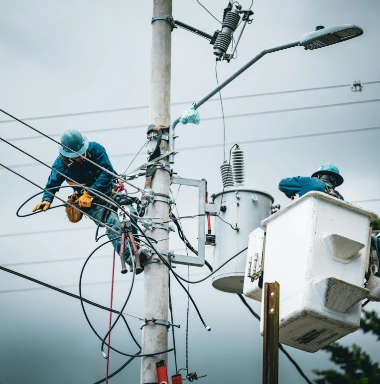 Two Men Focused On Maintenance Tasks On A Power Pole, Wearing Helmets And Harnesses For Safety